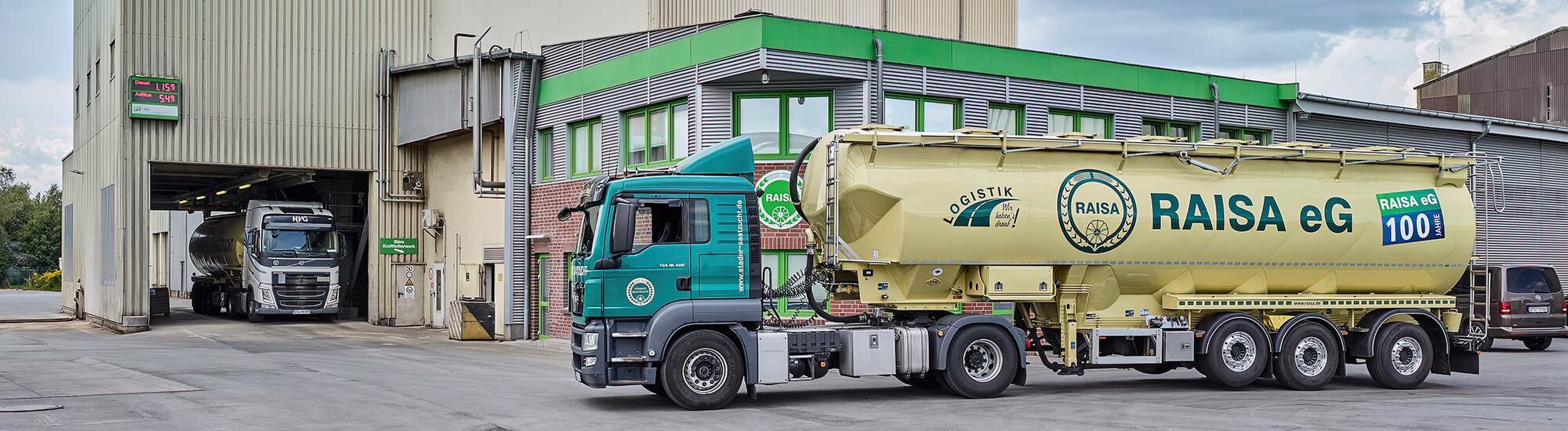 RAISA eG LKW mit gelbem Siloauflieger steht vor einer Industriehalle mit grünem Dach und Verladestation.