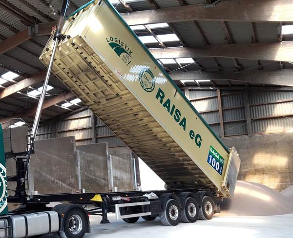 RAISA eG LKW kippt Schüttgut aus großem Auflieger in einer Lagerhalle mit offenen Dachfenstern ab.
