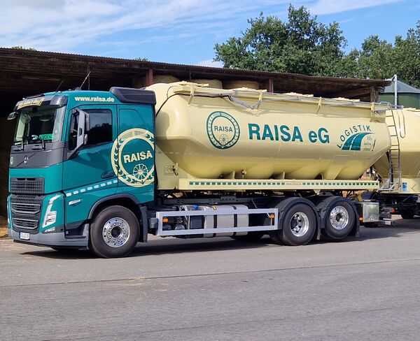 RAISA eG LKW mit Anhänger für Schüttgut steht auf einem Hof vor einer offenen Halle bei sonnigem Wetter.